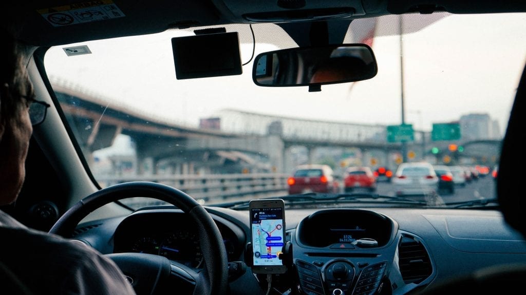 A man drives a car in traffic with the Uber app open on his smartphone. Taxi Cabs vs Uber & Lyft: Which Is Safer?